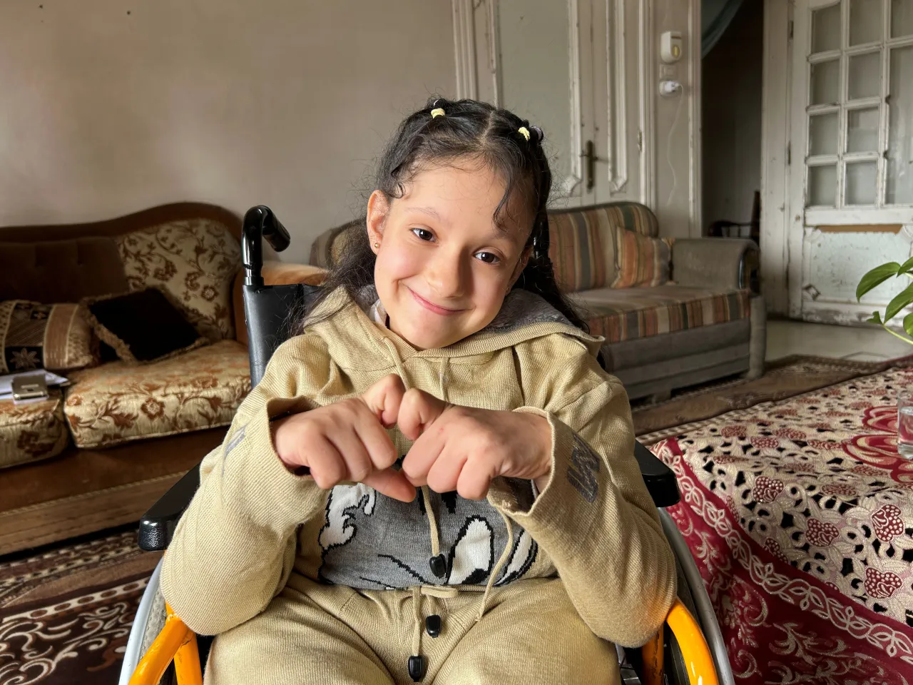 Sondus sits in a wheelchair inside a living room, smiling warmly at the camera with her hands clasped in front of her. She wears a light-colored sweatshirt and has her hair styled in small braids. Patterned rugs, a sofa, and an open doorway are visible in the background.