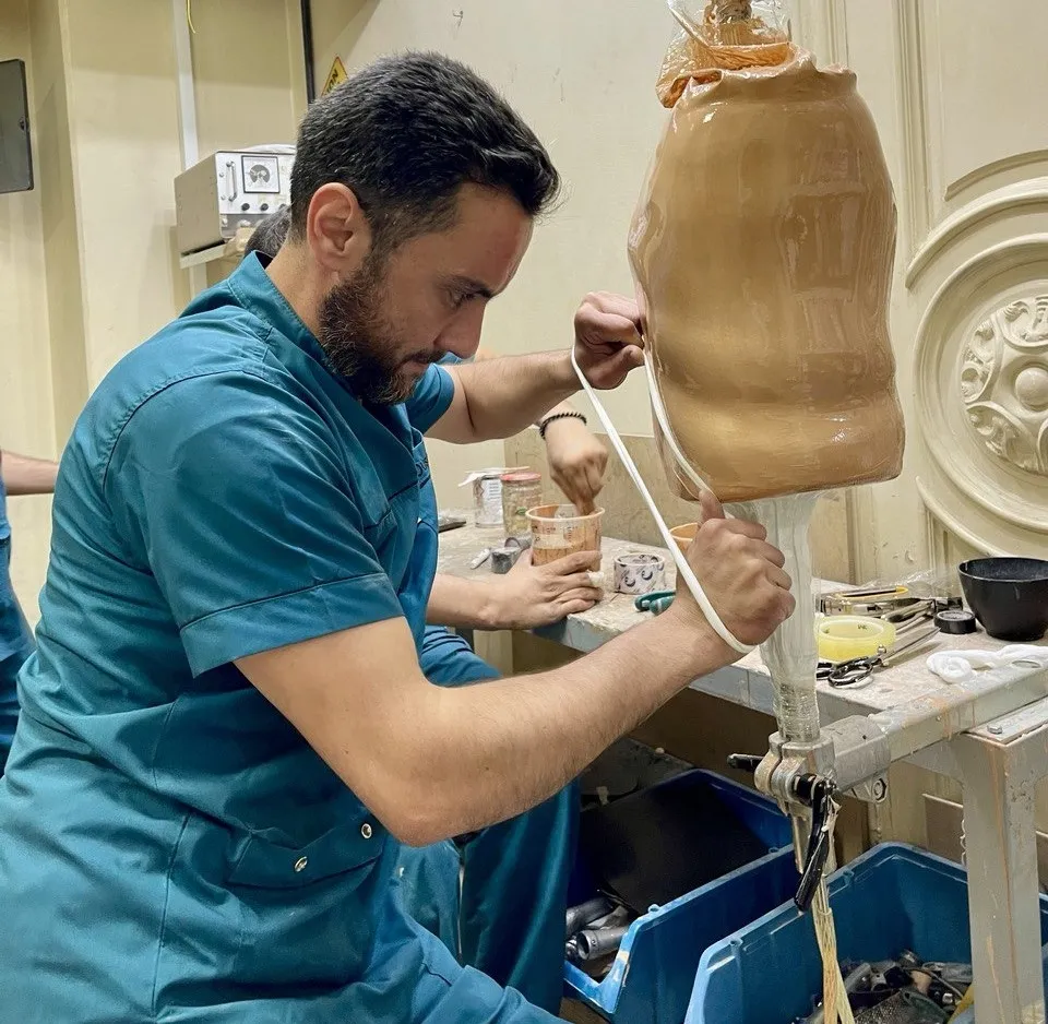 Ousama, a prosthetics technician in teal scrubs carefully adjusts a lower-limb prosthetic socket in a workshop. He works with focused attention, using tools on a workbench surrounded by equipment and materials.