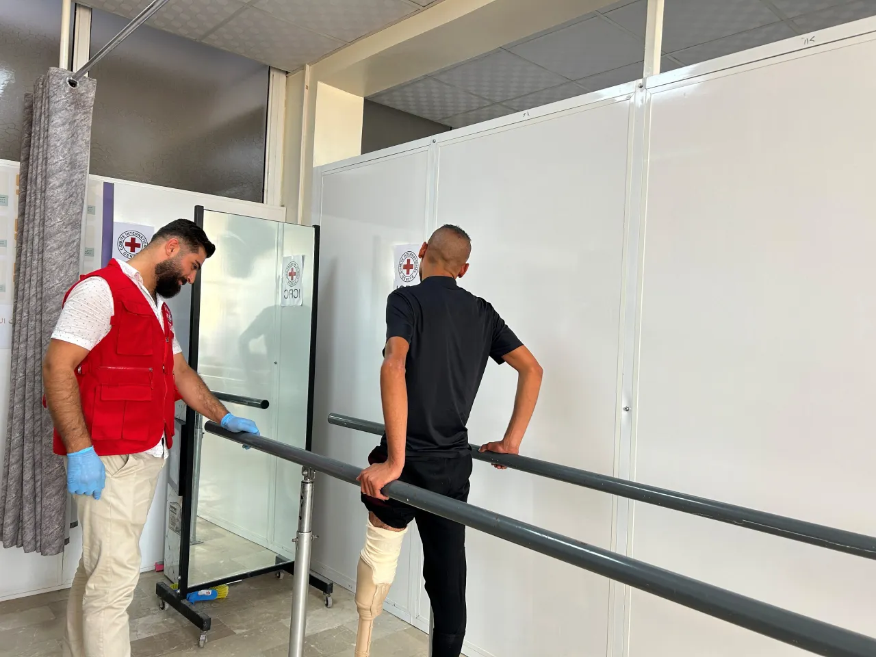 Khaled, a man with a lower-limb prosthesis, practices walking between parallel bars during a rehabilitation session. An ICRC staff member in a red vest stands beside him, observing and providing support in the bright training room.