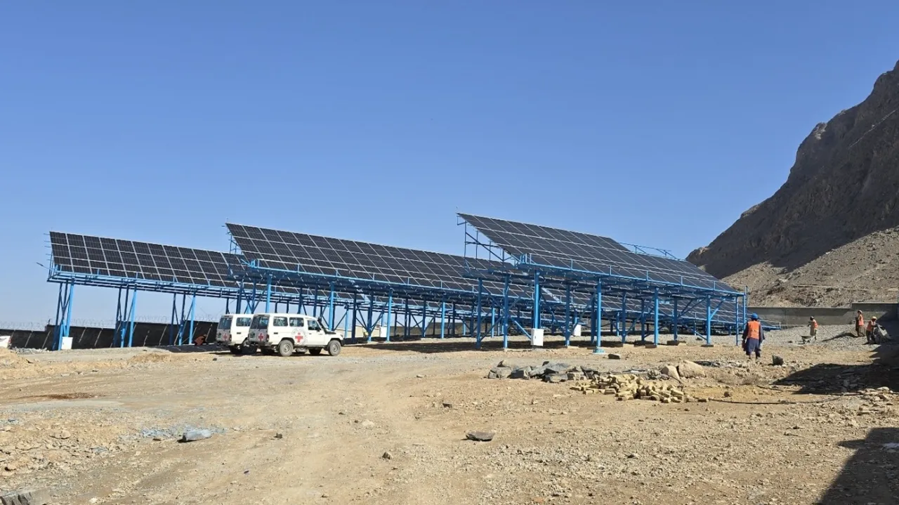 Rows of solar panels mounted on blue metal frames stand in a dry, mountainous landscape, with a few vehicles and people nearby under a clear sky.