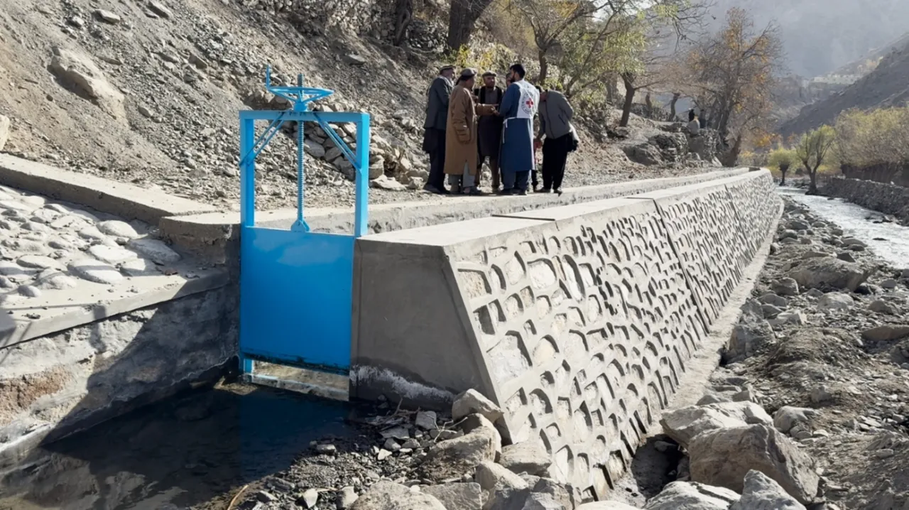 A small group of people stand beside a newly constructed concrete water channel with a blue metal gate in a rocky, mountainous area.
