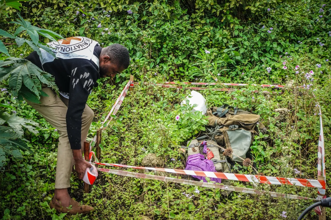 Un hombre coloca cinta de señalización alrededor de un área delimitada en medio de vegetación densa, donde se observan objetos en el suelo, indicando una zona potencialmente peligrosa.