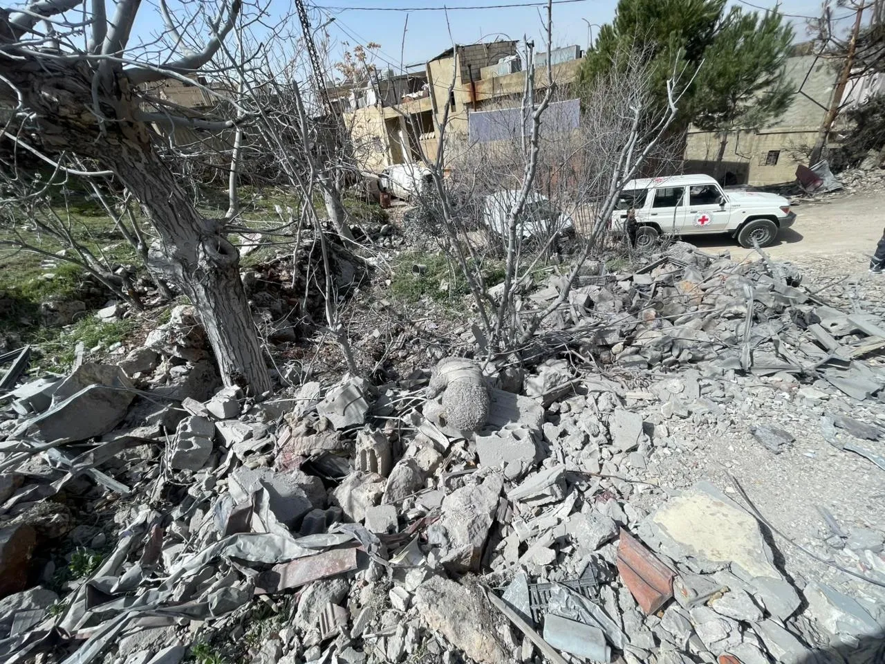 Escena de destrucción con escombros y restos esparcidos en el suelo. Entre los restos se ven árboles sin hojas, mientras que al fondo permanecen edificios intactos. Un vehículo blanco con el símbolo de CICR está estacionado cerca, indicando la presencia de servicios de emergencia o ayuda médica. 