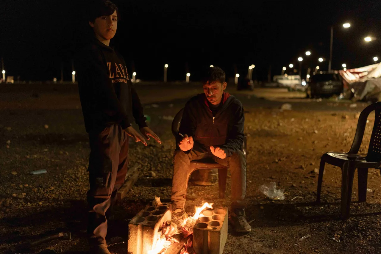 Two people gather around a small open fire at night, warming their hands beside makeshift seating in a displacement setting in Lebanon.