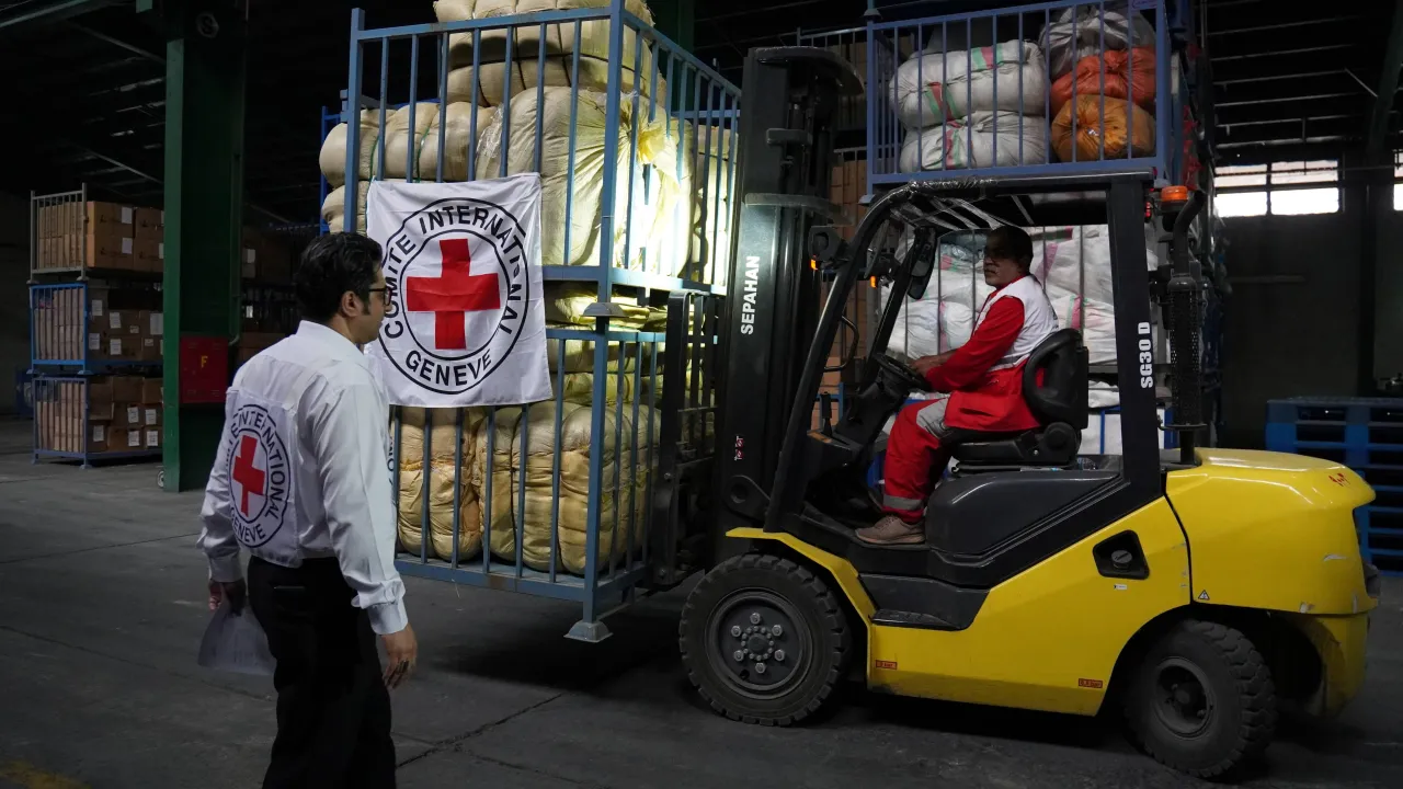 A forklift operated by a Red Crescent worker moves pallets of emergency relief supplies inside a warehouse in Iran, while another staff member oversees the operation.
