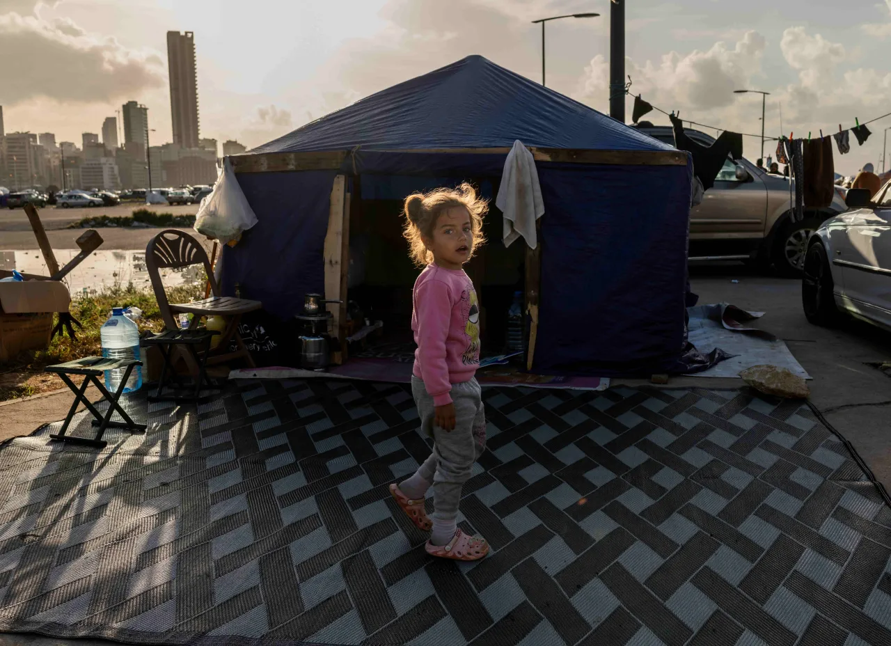 A young girl stands in front of a makeshift tent set up along a roadside, with city buildings visible in the background at sunset in Lebanon