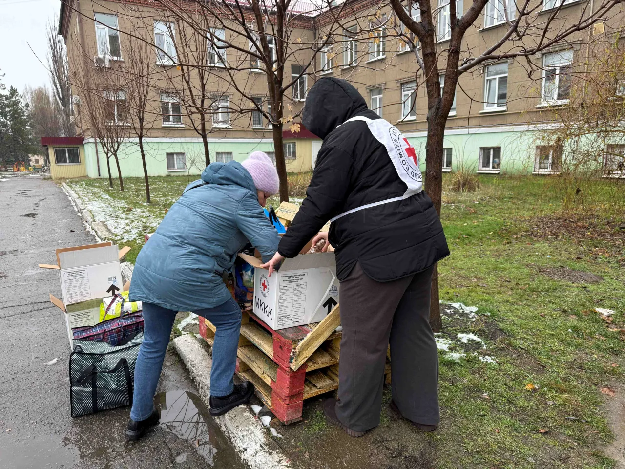 Two people stand outdoors beside stacked aid boxes marked with red cross symbols on wooden pallets, lifting or opening one box; a reusable shopping bag sits nearby, with apartment buildings and bare trees in the background.