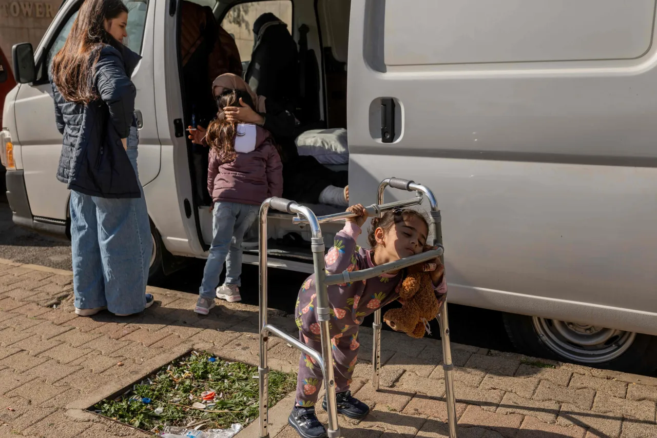 A young girl plays on her grandmother's walker outside a van, while others stand nearby and access belongings from inside