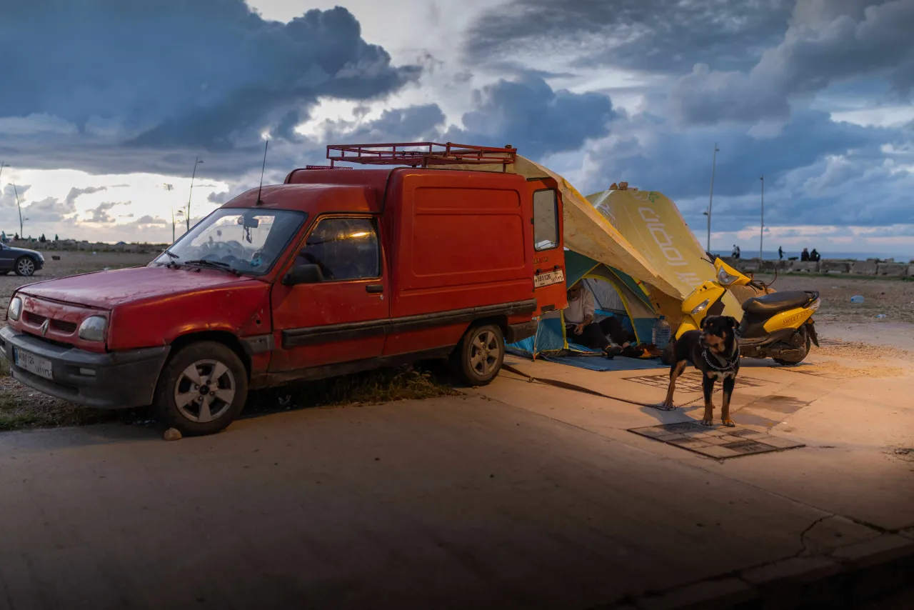 A red van is parked beside a tent set up along a roadside, with a person and a dog nearby, under a cloudy sky in Lebanon.