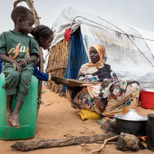 In a displacement camp in Sudan, a woman cooks over an open fire outside a makeshift shelter as two children watch.