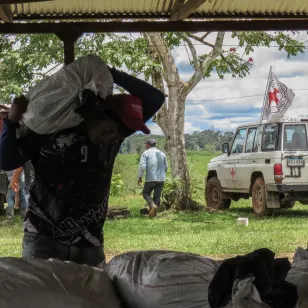 En primer plano, una persona carga un bulto con materiales, mientras al fondo se distingue un vehículo del CICR y varias personas en el entorno.
