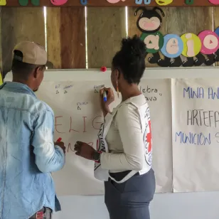 Una persona del CICR, de espaldas, llena una cartelera, mientras una persona de la comunidad, también de espaldas, observa con atención el contenido que se va construyendo