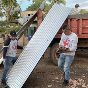 Dos personas del CICR se encuentran junto a un camión; una sostiene una teja, mientras la otra toma notas en un libro, en lo que parece una actividad de entrega o verificación de materiales.