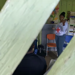 nmarcada entre dos palos en primer plano, se observa al fondo a una persona del CICR facilitando un taller o dirigiéndose a un grupo. La imagen crea un efecto natural de encuadre que centra la atención en la escena.