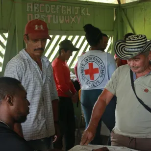 En un salón comunitario, varias personas están reunidas. En primer plano, tres observan atentamente lo dispuesto sobre una mesa, mientras al fondo, una persona del CICR conversa con otro grupo.