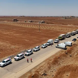 A long humanitarian aid convoy stretches along a rural road in Sweida, Syria, in July 2025. The convoy includes dozens of white vehicles marked with the Red Cross and Red Crescent emblems, along with large trucks carrying relief supplies. The road runs through a dry, reddish-brown landscape with sparse vegetation and scattered buildings in the distance. Aid workers in red uniforms walk near the vehicles under a clear blue sky.