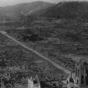 A black and white photograph showing the devastated landscape of Hiroshima after the nuclear bombing in August 1945. The area is flattened and burned, with only a few skeletal buildings and bare trees remaining. A straight dirt road runs through the center, surrounded by rubble and destruction, with mountains visible in the background.