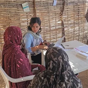 An ICRC worker sits at a small table inside a shelter made of woven branches, speaking with two women who sit opposite her. The women wear patterned headscarves, and the aid worker uses a laptop while taking notes. Sunlight filters through the gaps in the shelter walls.