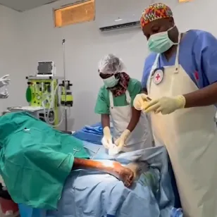 A medical team performs surgery in a clean, bright operating room. Three health workers in scrubs, masks, and gloves stand around a patient lying on an operating table, with one surgeon focused on the procedure under bright surgical lighting. Monitors and medical equipment are visible in the background.