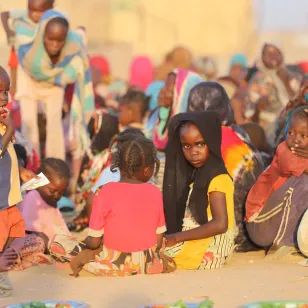A group of displaced families sits together on sandy ground in Tawila, Sudan. Several women and children gather closely, some looking toward the camera. In the foreground, a young boy stands holding a piece of paper while another child in a headscarf sits among the group. The scene reflects the difficult conditions people are facing after fleeing violence.