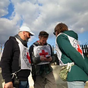 Three ICRC staff members wearing Red Cross vests stand together reviewing notes outside a fenced area topped with barbed wire in Lebanon during humanitarian operations amid escalating hostilities.