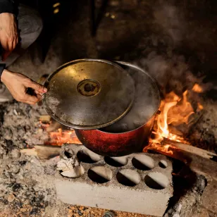 Hands lift the lid from a pot cooking over an open fire, with makeshift supports and burning wood in a displacement setting in Lebanon.