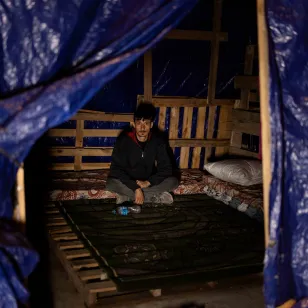 Haitham sits on a mattress inside a makeshift shelter framed by blue tarpaulin, with simple bedding and wooden pallets forming the structure in Lebanon.