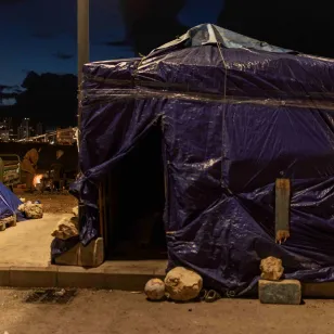 A makeshift shelter covered in plastic sheeting stands along a roadside at night, with nearby tents and city lights visible in the background in Lebanon.