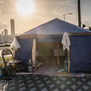A makeshift tent stands along a roadside at sunset, with chairs, water containers and personal belongings arranged outside