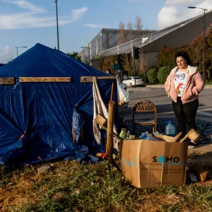 Farah stands beside a makeshift shelter set up along a roadside, with household items and supplies arranged outside