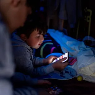 A child lies on bedding inside a tent, illuminated by the glow of a mobile phone
