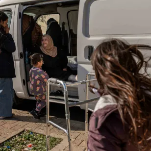 Saada sits at the back of a van where she lives with her extended family, while a child stands nearby and another person speaks with her outside.