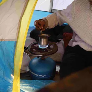 Inside a tent, Zaher prepares coffee on a small gas stove, with basic cooking items arranged on the ground in a displacement setting in Lebanon.