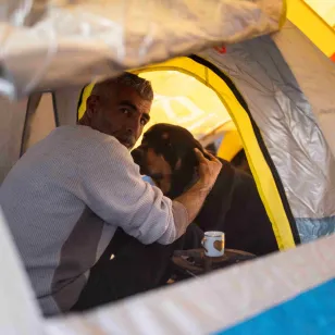 Zaher sits inside a small tent, holding his dog close, with a cup placed beside him, in a displacement setting in Lebanon.