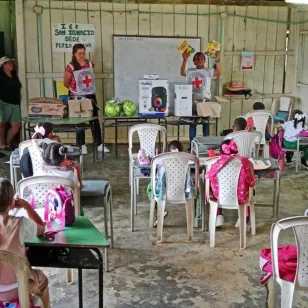 Salón de clases con niños sentados mientras personas adultas se encuentran al frente.