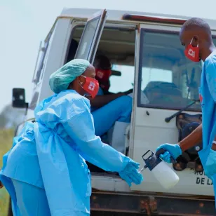 Red Cross volunteers in blue protective clothing handle disinfection equipment beside a vehicle during Ebola response operations in the Democratic Republic of the Congo.