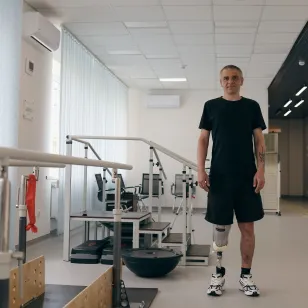 A man with two lower-leg prosthetic limbs stands in a rehabilitation centre beside parallel bars used for gait training. Exercise equipment is visible in the background of the bright therapy room.