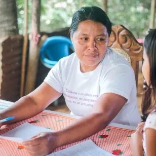 Una mujer está sentada frente a una mesa, con un bolígrafo y una hoja de papel en las manos, mientras conversa con una niña sentada a su lado. Sobre la mesa hay varias hojas, entre ellas una fotografía.