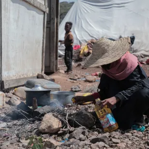 A person cooks over a small outdoor fire among rubble and makeshift shelters.