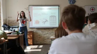 ICRC delelgate talking to students seated at desks in a classroom with information about explosive ordanances on a board behind her