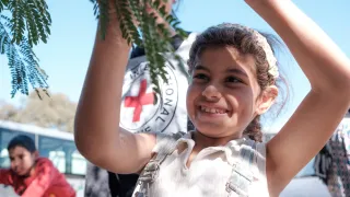 Batoul plays with a tree branch, smiling, with blue sky and a red cross in the background