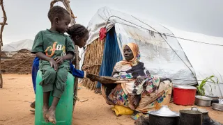 In a displacement camp in Sudan, a woman cooks over an open fire outside a makeshift shelter as two children watch.