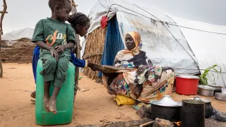In a displacement camp in Sudan, a woman cooks over an open fire outside a makeshift shelter as two children watch.