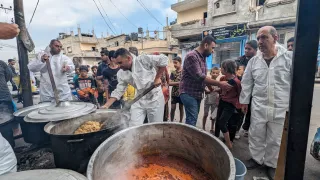 Workers in white overalls stir large vats of food in the community kitchen, people and buildings in the background