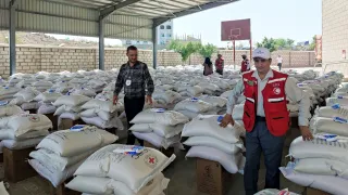 Two humanitarian workers (one from the ICRC and another one from the Yemeni Red Crescent) stand near each other surrounded by many piles of bags and boxes of food items