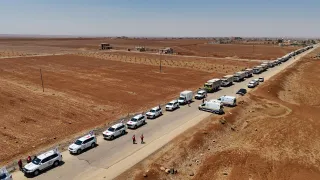 A long humanitarian aid convoy stretches along a rural road in Sweida, Syria, in July 2025. The convoy includes dozens of white vehicles marked with the Red Cross and Red Crescent emblems, along with large trucks carrying relief supplies. The road runs through a dry, reddish-brown landscape with sparse vegetation and scattered buildings in the distance. Aid workers in red uniforms walk near the vehicles under a clear blue sky.