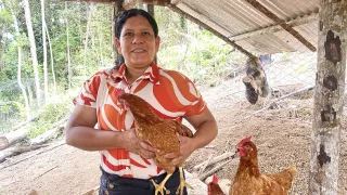 Mujer dentro de un gallinero, sostiene una gallina entre sus manos y sonríe a la cámara.