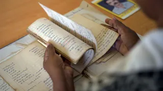 A person flips through the pages of an old, handwritten notebook. Several other aged notebooks lie on the table, along with a photograph of a man placed in the background.
