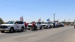 A convoy of white vehicles marked with Red Cross emblems and flags parked on a road under a clear blue sky, with humanitarian workers standing beside them.