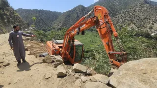 A man wearing a Red Crescent emblem points towards an excavator off a dirt road on a steep hillside in a mountainous area of Afghanistan, following the earthquake.